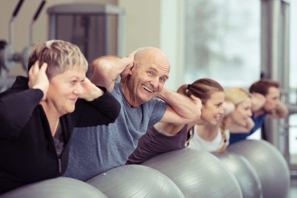 Elderly couple doing pilates class at the gym with a group of diverse younger people balancing on the gym ball with raised arms to tone their muscles in an active retirement concept-3